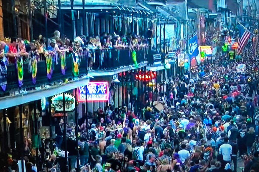 Crowd of revelers in vibrant costumes and masks celebrating Mardi Gras on a bustling New Orleans street, with colorful floats and decorations.