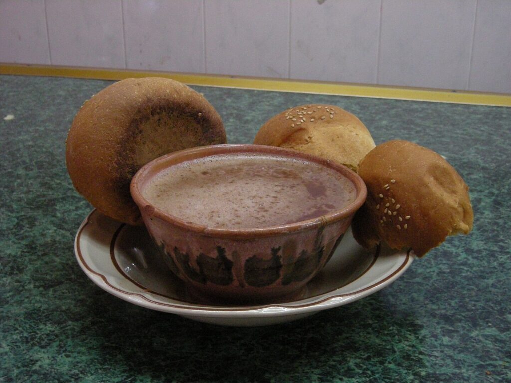 A cup of Oaxacan-style hot chocolate served in a traditional handleless clay mug, accompanied by three pieces of pan de yema (egg-yolk bread).